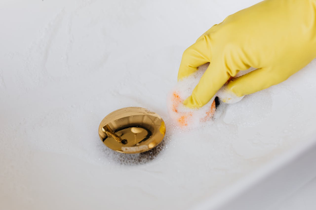 Why Post-Placement Support Matters When Hiring a Maid in Singapore Close-up of a hand in a glove cleaning a foam-covered gold drain in a sink.