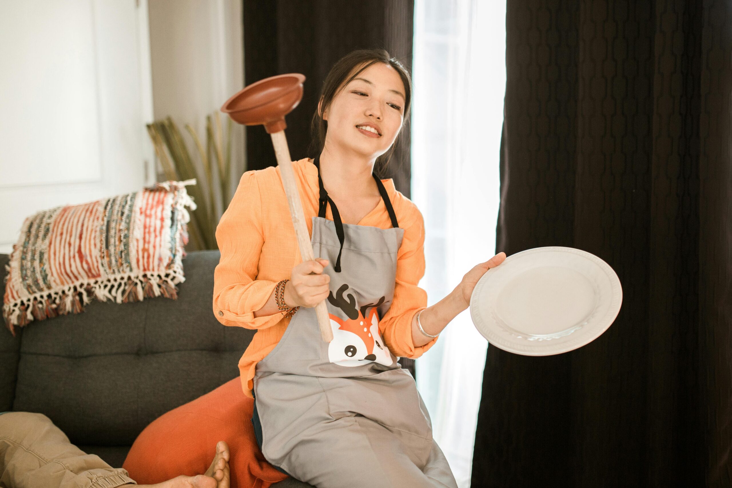 How to Hire the Right Maid in Singapore: A Complete 2025 Guide A woman with an apron playfully holds a plunger and plate indoors, showcasing a lighthearted home scene.